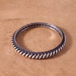 A silver-toned twisted rope bangle bracelet resting on a textured brown surface.