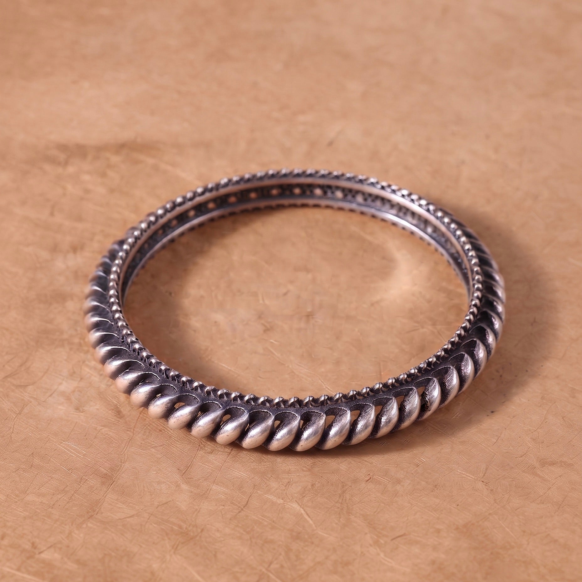 A silver-toned twisted rope bangle bracelet resting on a textured brown surface.
