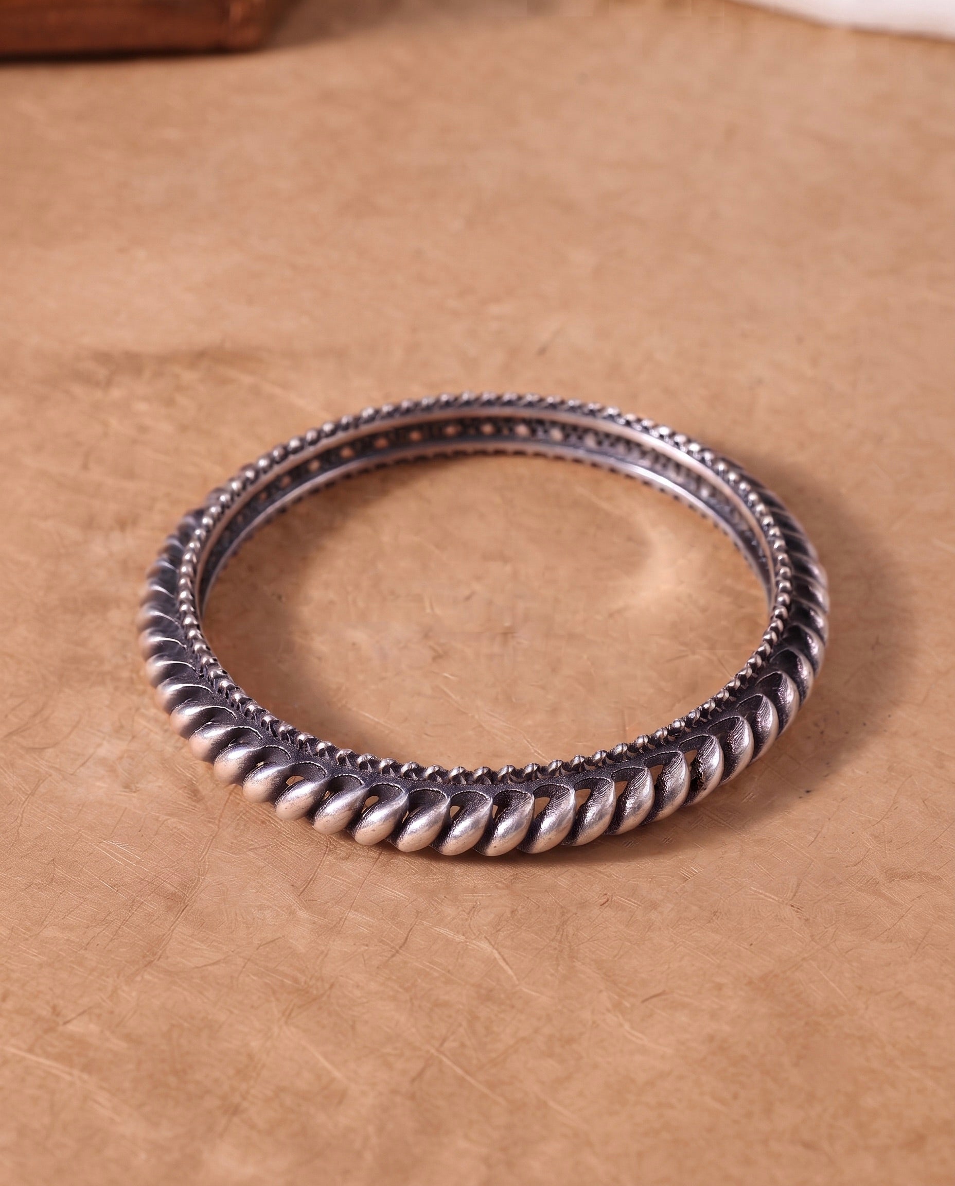 A silver-toned twisted rope bangle bracelet resting on a textured brown surface.