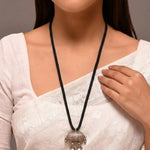 Woman wearing a white saree with a black necklace featuring a large pendant.